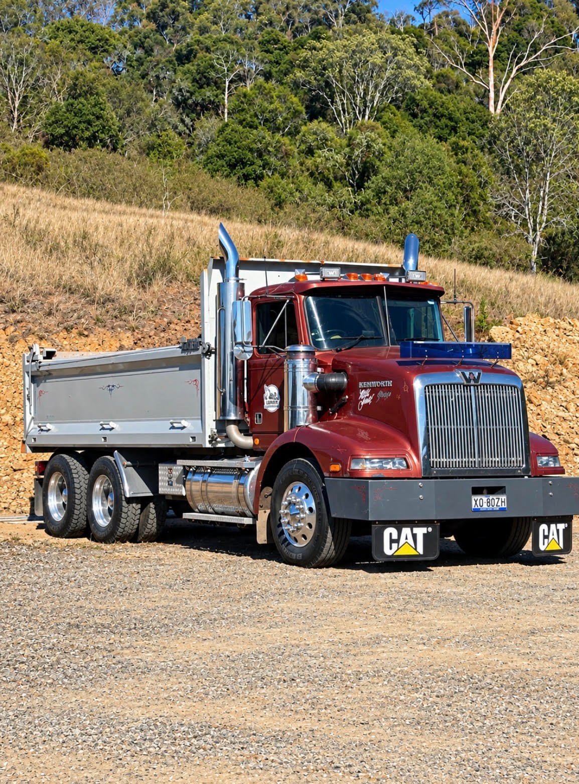 Red and blue Caterpillar dump truck with chrome accents parked on gravel road with trees and dry grass in background